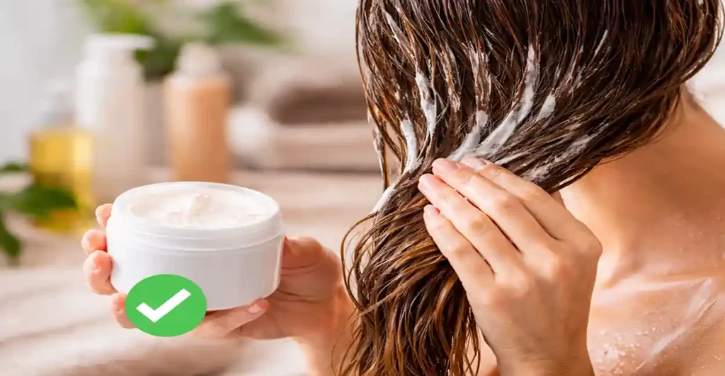 Imagem realista em formato 16:9 mostra uma mulher aplicando uma máscara capilar cremosa nos fios úmidos após a lavagem. O cabelo aparece alinhado e bem distribuído, enquanto a textura do produto é visível ao longo do comprimento. Ao lado, um pote aberto de creme reforça a ideia de tratamento capilar. A cena transmite um momento de cuidado e hidratação profunda, com iluminação suave e ambiente clean ao fundo.