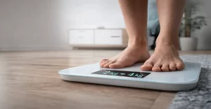 Imagem em formato 16:9 mostrando um close nos pés de uma pessoa posicionados sobre uma balança digital moderna. O ambiente é doméstico, com fundo levemente desfocado e iluminação suave, transmitindo sensação de rotina e controle de peso. A composição visual reforça a ideia de monitoramento do emagrecimento de forma segura e consciente, sem elementos distrativos ou textos na imagem.